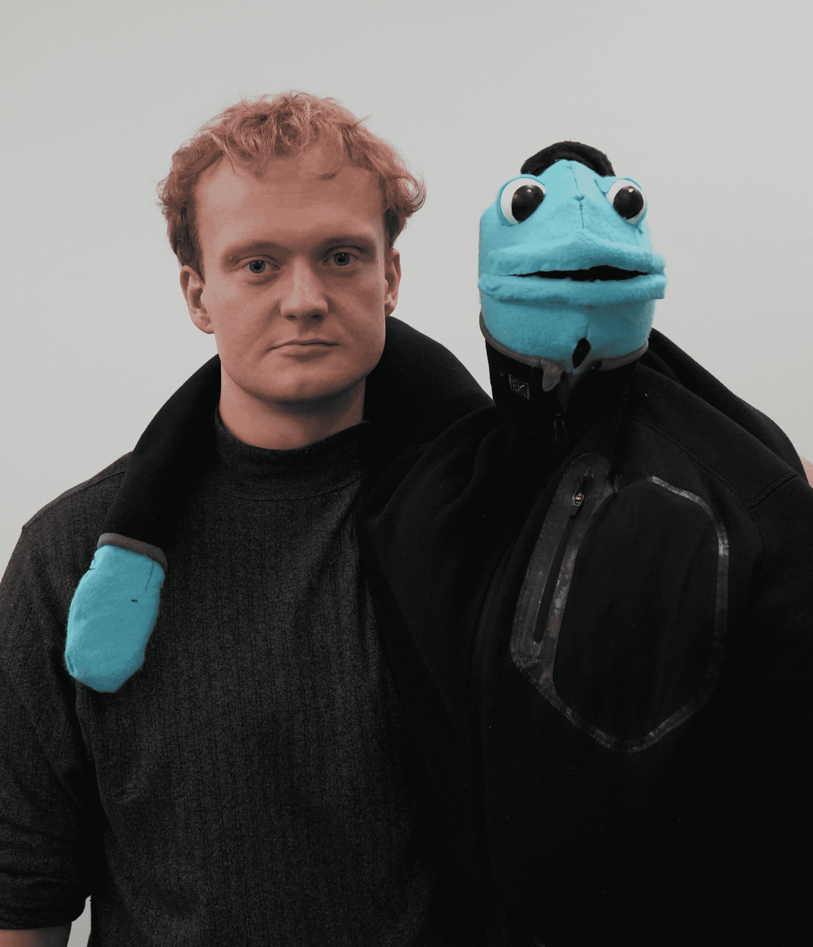 A blonde man in a tutrleneck sweater with a blue puppet on his hand.
