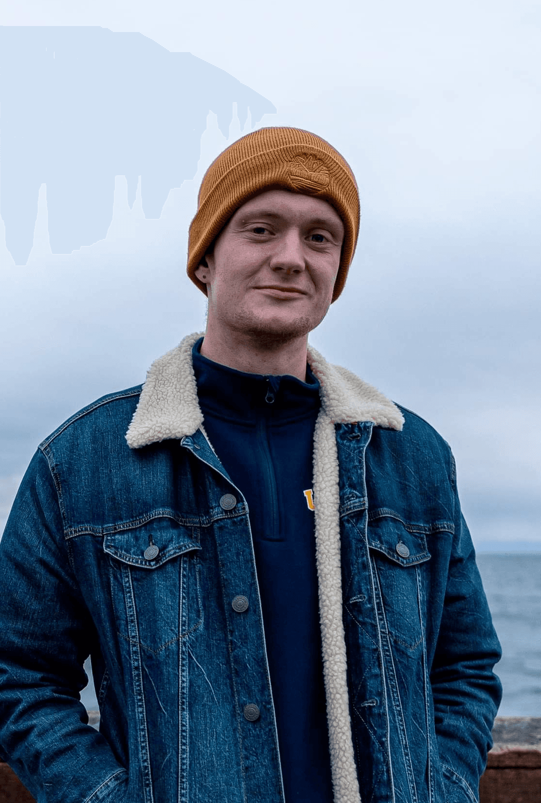 A tall Latvian-American man wearing an orange beanie standing in-front of an ocean view. His hands are tucked into the pockets of his jacket.